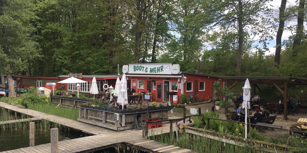 Bild, das die Steganlage von Boot & Mehr am Kleinen Pälitzsee zeigt, ein roter Flachbau mit blumengeschmücktem Biergarten davor.