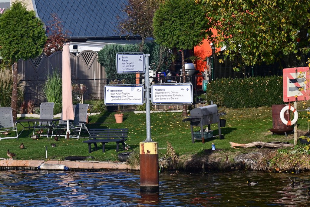 Zu sehen ist ein Wegweiser der mit drei Tafeln den Weg nach Köpenick, Neukölln/Potsdamm und Berlin-Mitte/Kreuzberg zeigt. In Hintergrund ein Ufergarten mit Grill und Sitzecke.