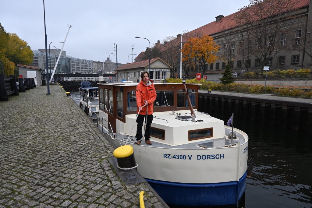 Zu sehen ist ein Hausboot beim Schleusenvorgang, auf dem Vordeck steht der jugendliche Sohn der eine Festmacherleine um einen Poller führt.