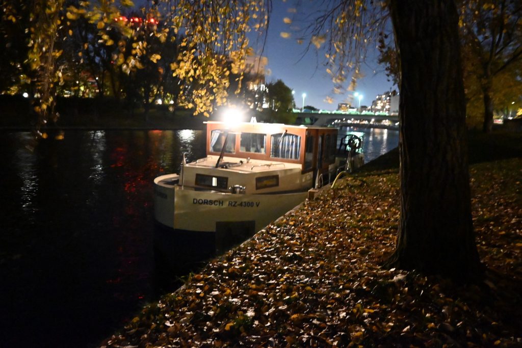 Zu sehen ist ein Hausboot im fast ganz verschwunden Tageslicht, es liegt an einer Spundwand am mit herbstlichen Blättern bedecktem Ufer, im Hintergrund sieht man die Lichter einer Brücke und der Stadt Berlin.