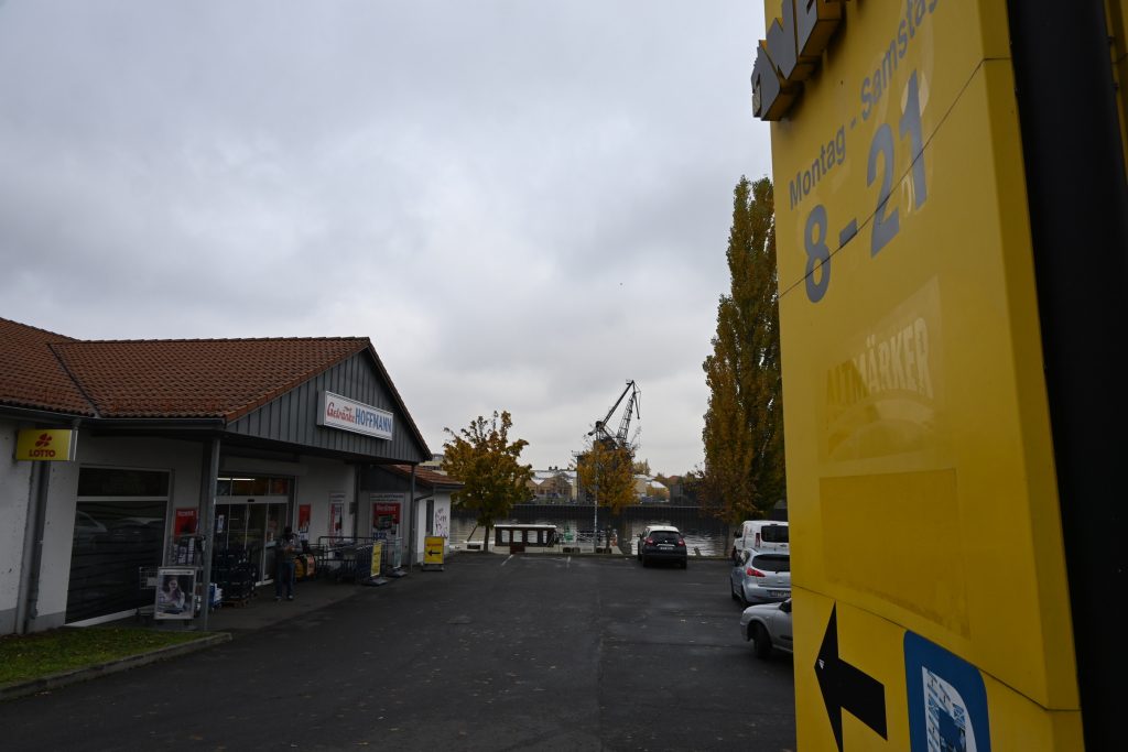 Zu sehen ist ein Supermarktparkplatz zwischen zwei Einkaufsmärkten. Im Hintergrund ist das Deckshaus einer im Wasser liegenden Kormoran zu sehen.