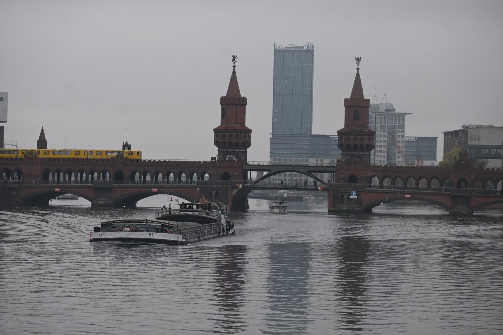 Zu sehen ist ein Binnenfrachtschiff in Fahrt im Hintergrund die Oberbaumbrücke mit den charakteristischen Türmchen und einer gelben U-Bahn.
