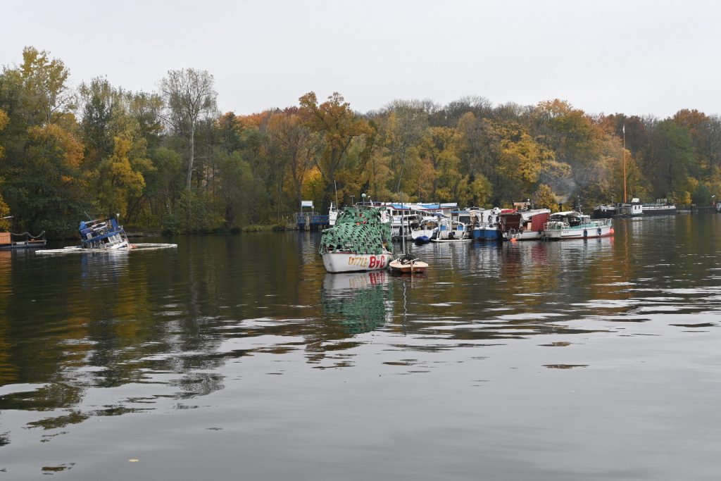 Zu sehen sind eine Anzahl alter und sehr alter Boote, ein Boot liegt halb gesunden und von einer Ölsperre umgeben etwas abseits.