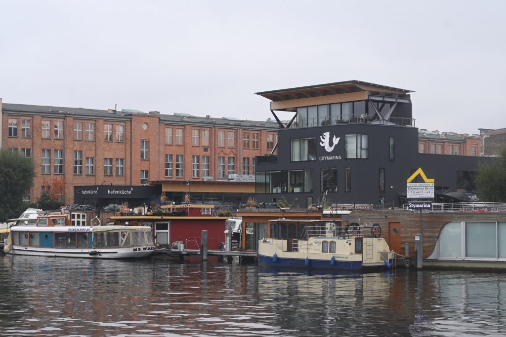 Zu sehen ist der Hafen der Citymarina Berlin Rummelsburg, im Hintergrund teilweise moderne Gebäude, teilweise alte Industriearchitektur mit Backsteingebäuden.