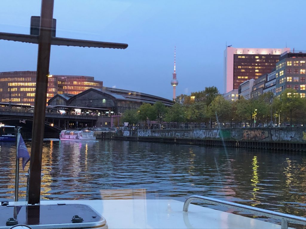 Zu sehen ist der Blick vom Schiffsbug auf Wasser im Hintergrund ist der Bahnhof Friedrichstraße zu sehen. Alles zur blauen Stunde, also mit Abendstimmung.