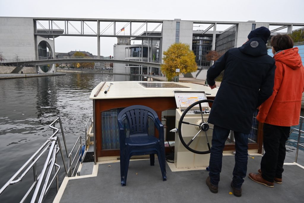 Zu sehen ist das Achterdeck eines Hausbootes mit Fahrstand Vater und Sohn am Stuern, im Hintergrund Regierungsgebäude.
